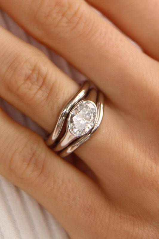 A close-up of a hand wearing a white gold signet with a clear oval diamond -cut gemstone, resting on a white ribbed fabric surface.