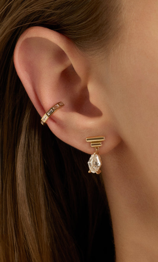 A close-up of a model's ear showcasing a gold ear cuff with small clear stones and a matching gold drop earring with a teardrop-shaped clear crystal.