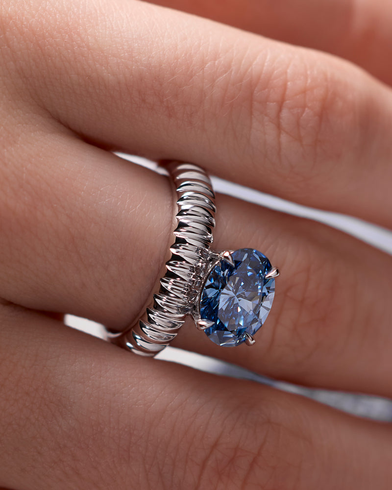 Close-up of a hand wearing a signature architectural LEIL rings in 18k white gold with a large blue lab diamond on a neutral background