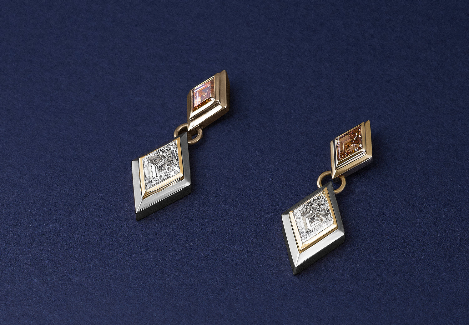 A pair of geometric double bezel drop earrings in yellow gold and white gold double bezel, featuring lozenge cut brown and white lab diamonds, displayed on a dark navy blue background.