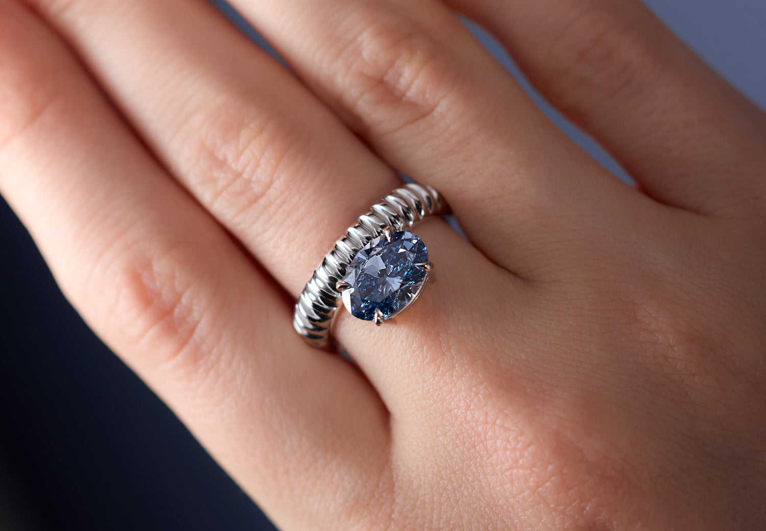 A close-up of a hand wearing a signature Leil Auri architectural solid white gold ring with a large oval lab grown blue diamond. 