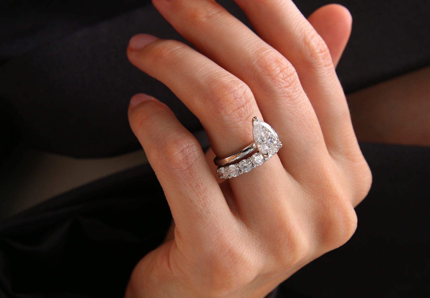 A close-up of a hand wearing a white gold Leil engagement ring with a pear-cut lab-grown diamond and a matching band with round brilliant-cut diamonds, set against a dark background.