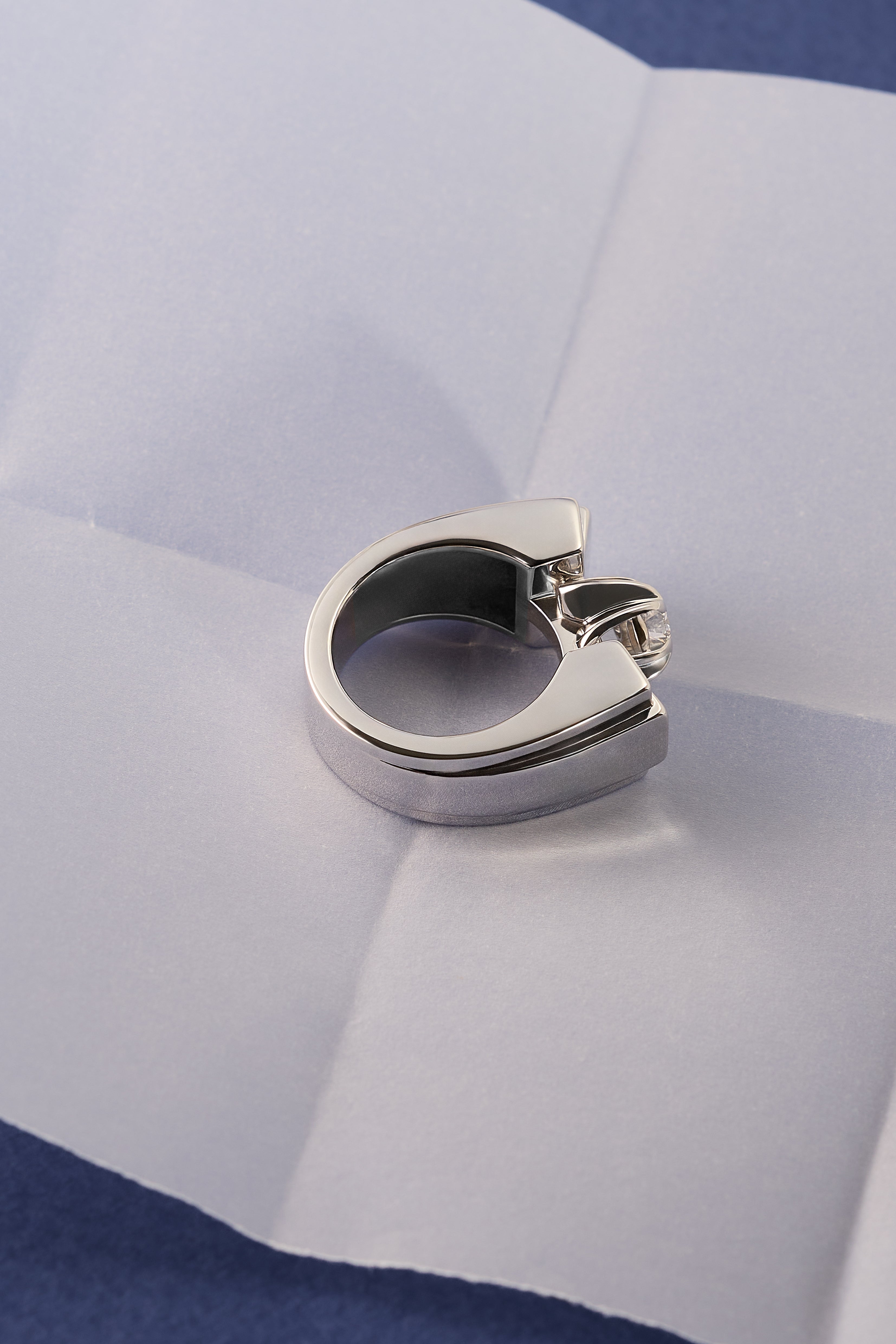 A polished platinum and diamond ring with a unique band design, resting on a white sheet of paper with a blue background visible in the corners.