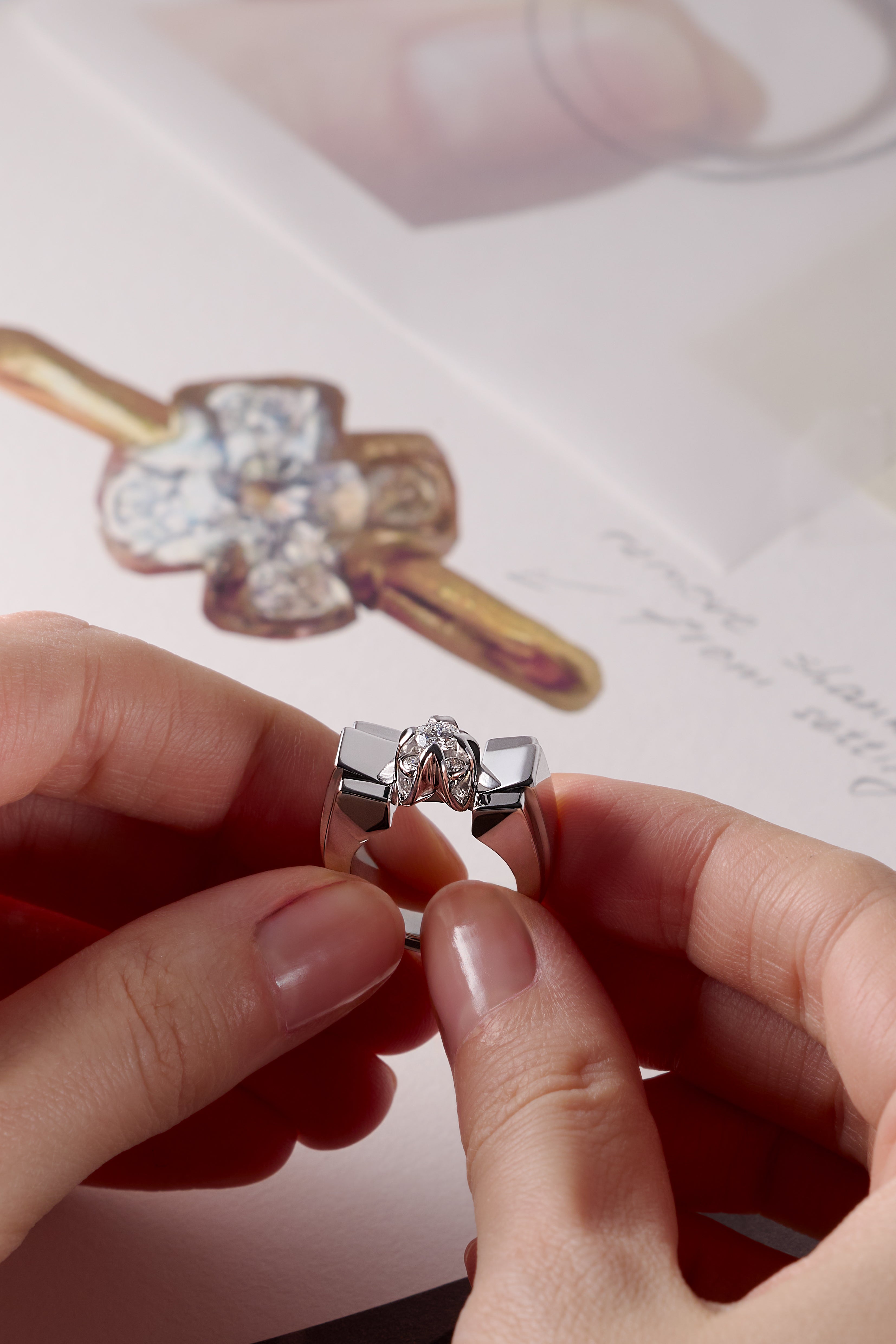 Person holding a bespoke platinum Leil ring with a diamond, with a blurred background of sketches detailing the bespoke process.