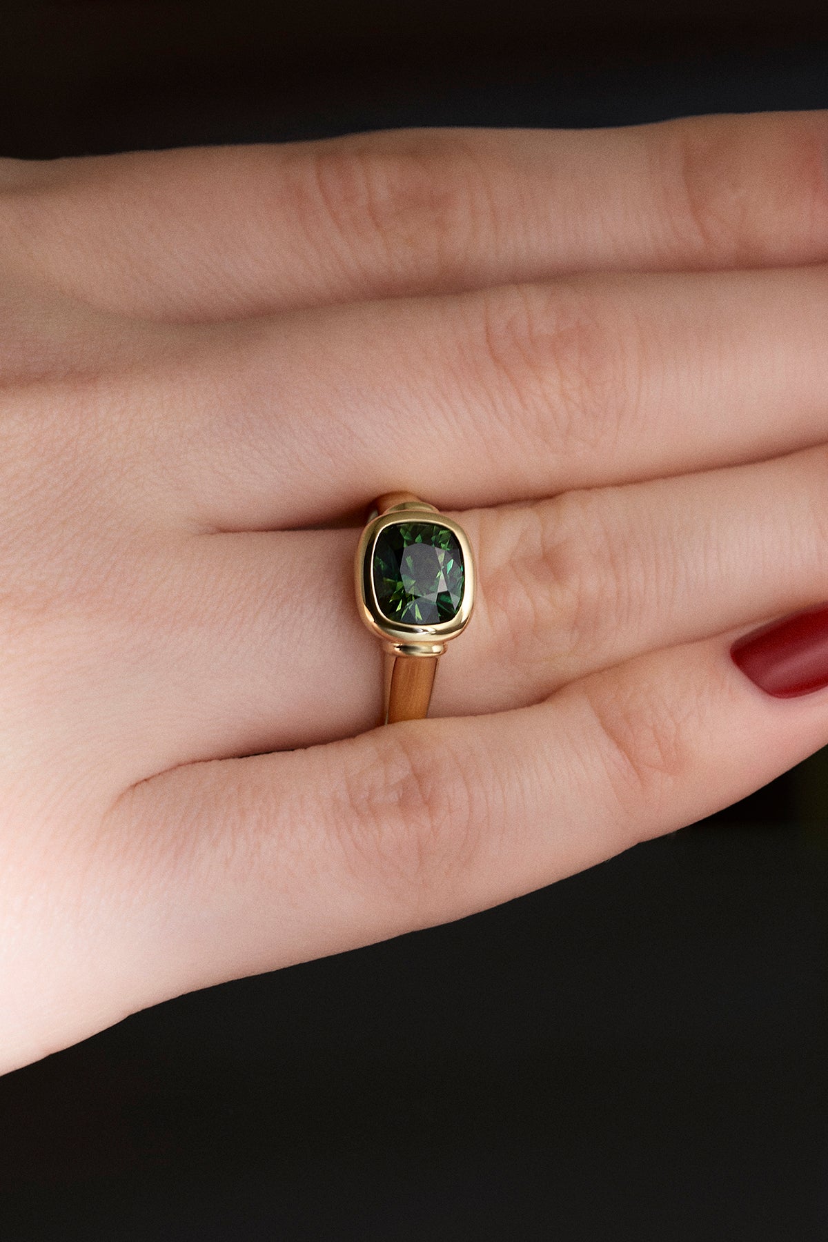 A yellow gold Leil Ionia ring with a cushion-cut green diamond, shown worn on a person's finger against a dark background.