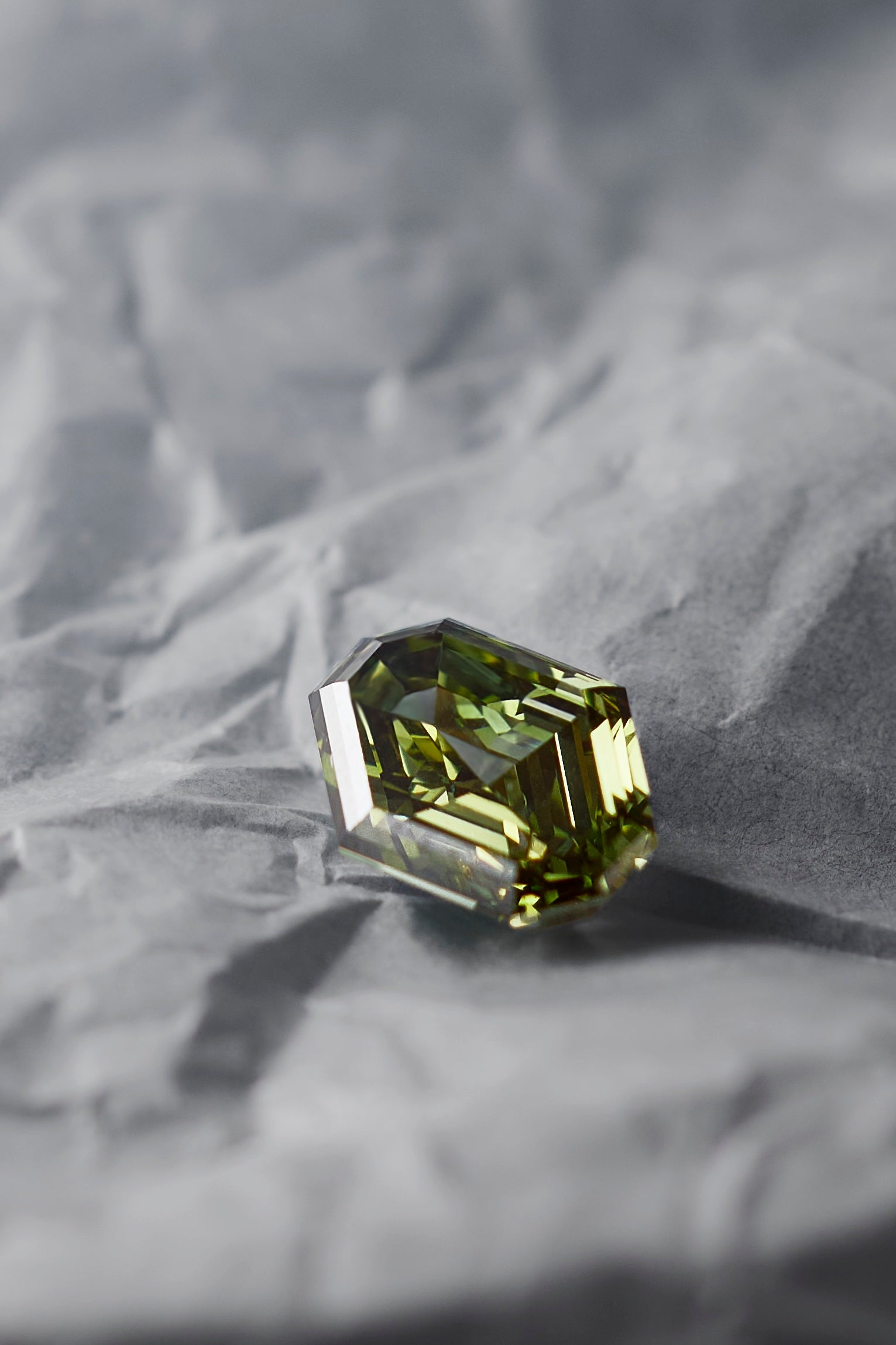 A 2.5 carat olive-green shield-cut lab grown diamond resting on a textured gray crumpled paper background.