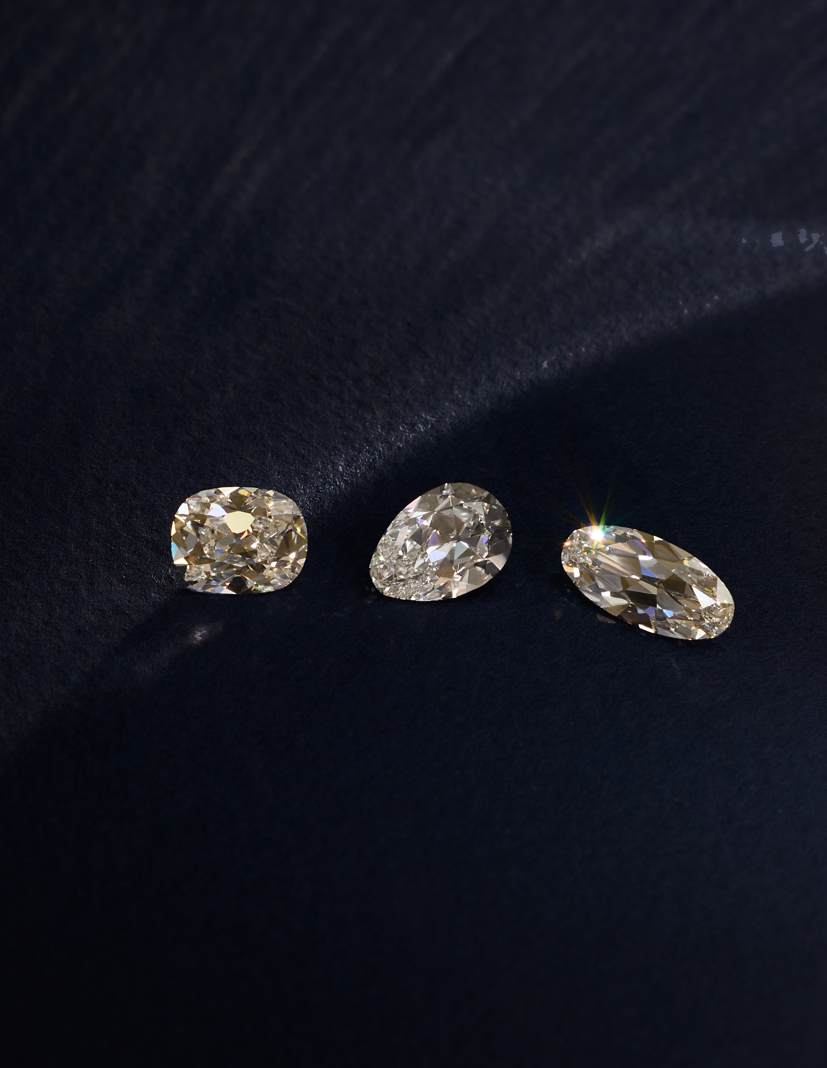 Three loose old mine cut diamonds in cushion, pear, and marquise shapes with a warm champagne color, arranged on a dark navy blue textured fabric background.